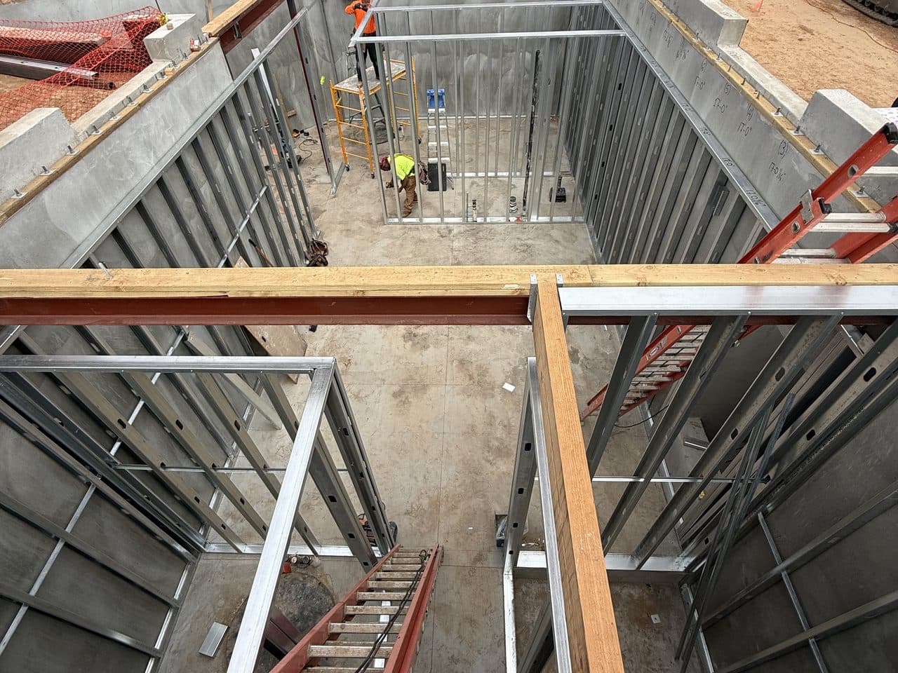 Concrete substructure walls and light gauge steel framing going up on a below-grade level, with crews working in the pit.