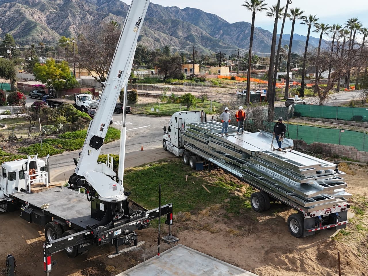 A boom crane and flatbed delivering panelized light gauge steel to a foothill jobsite, with the San Gabriel mountains behind.